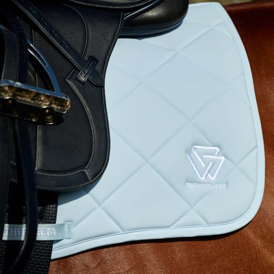 A close-up of a light blue quilted equestrian saddle pad with a white embroidered logo, shown under a black leather saddle on a brown leather surface.